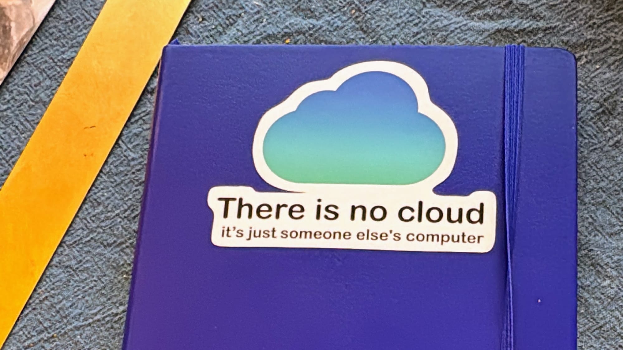 A notebook on a desk with a sticker on it, which says "There is no cloud. It's just someone else's computer"